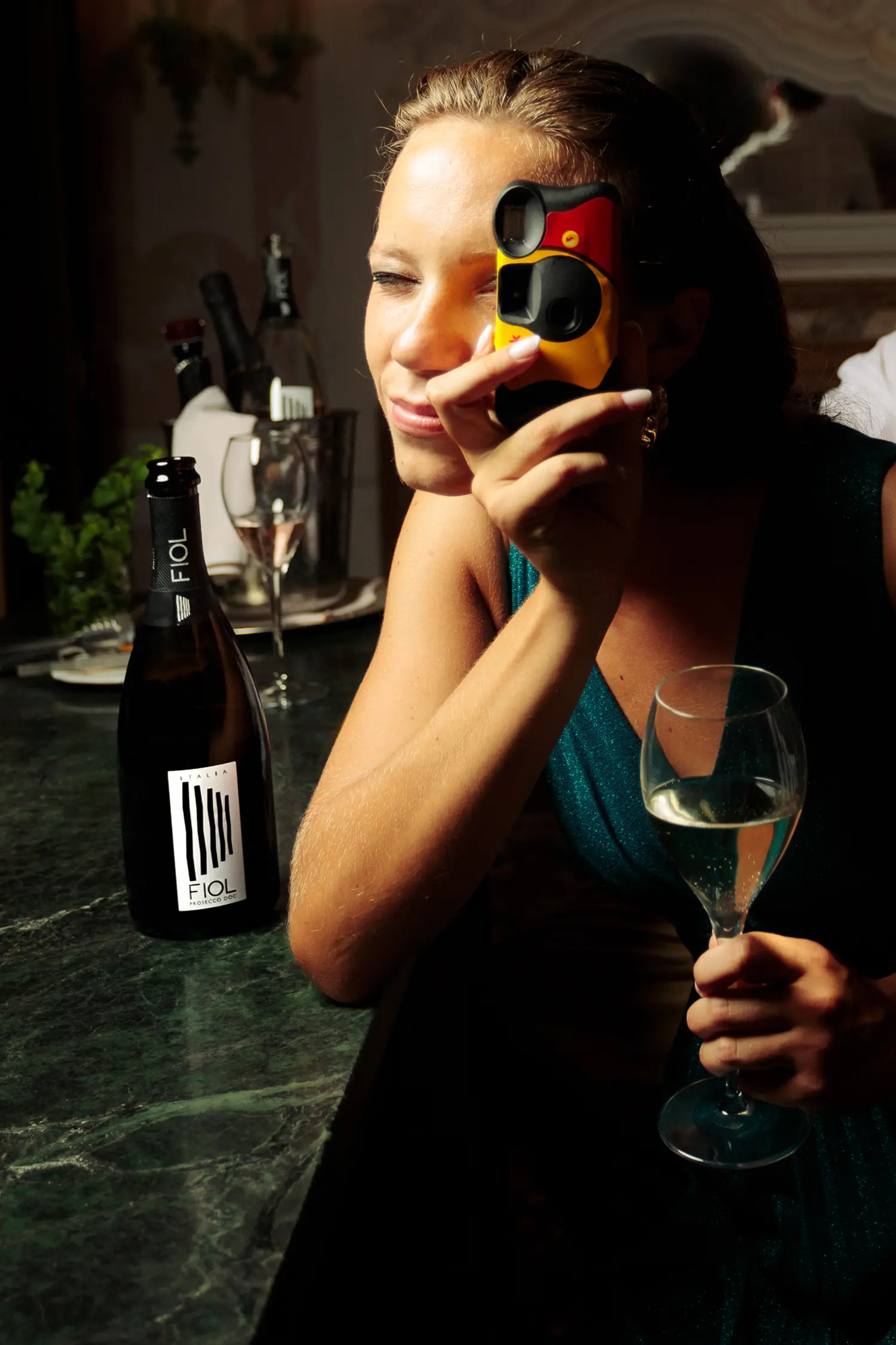 Una ragazza elegantemente vestita, durante una festa, tiene in mano un calice di Prosecco mentre scatta una foto con una macchina fotografica analogica.