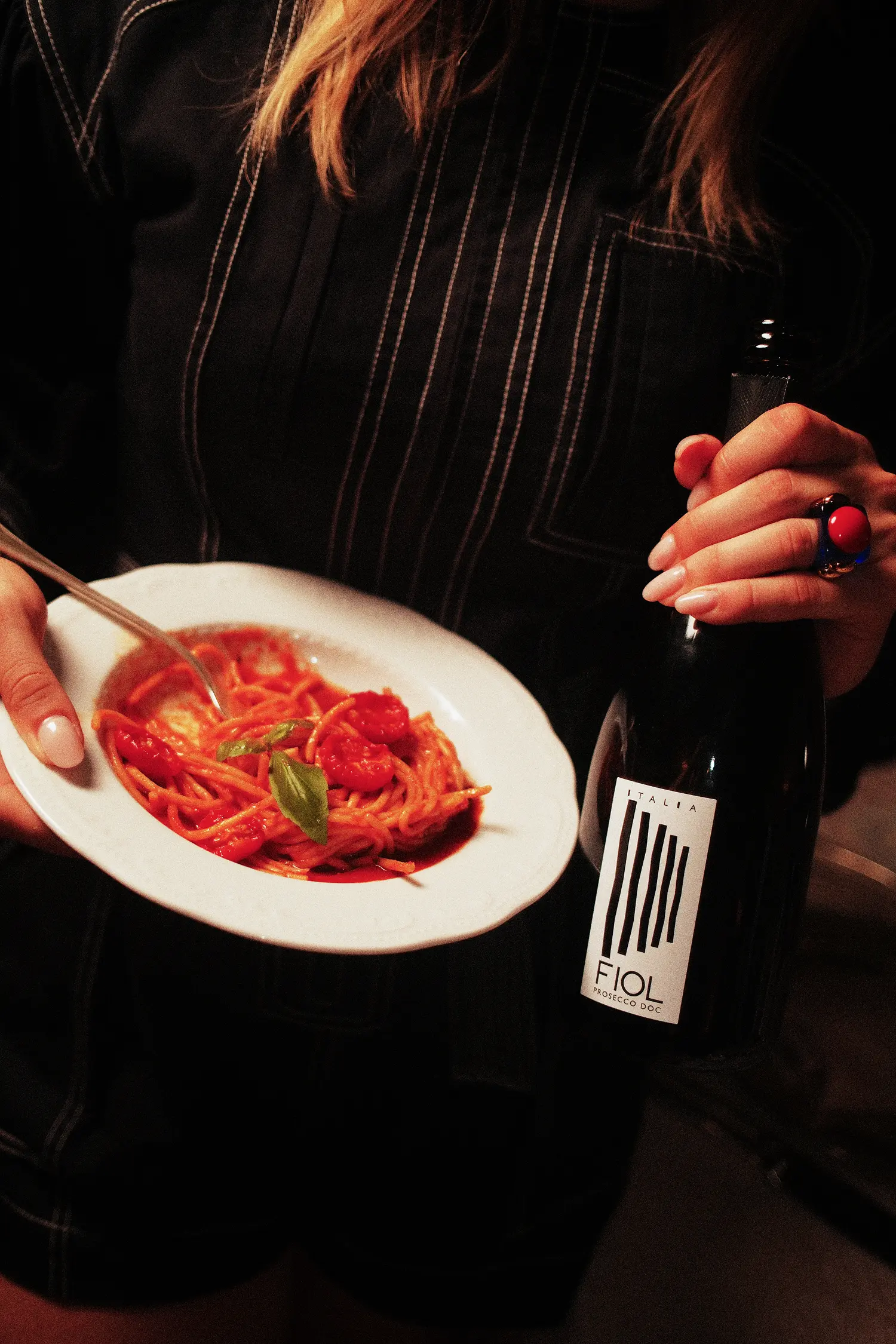 Una ragazza fotografata in un contesto serale, regge tra le mani un piatto di spaghetti al pomodoro e una bottiglia di prosecco Fiol